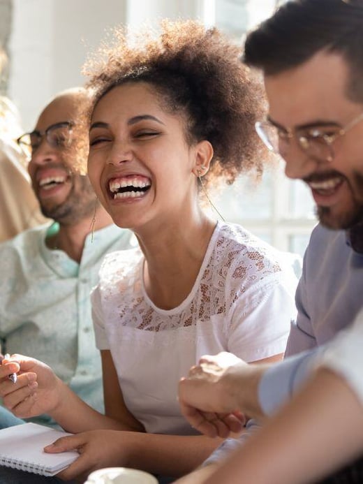 A group of diverse colleagues sitting together and laughing, showcasing a joyful and collaborative environment. The focus is on a woman with curly hair in a white top, smiling brightly.