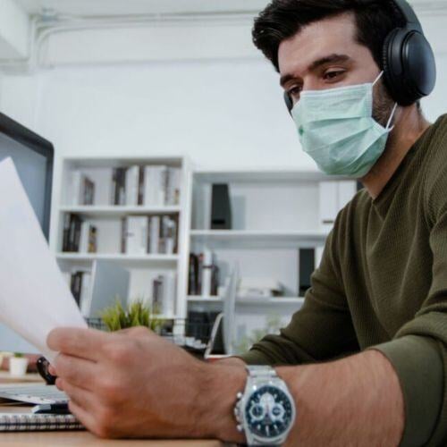 Image shows person wearing a face covering and headphones whilst reading a document. They are wearing a green jumper with the sleeves rolled up.