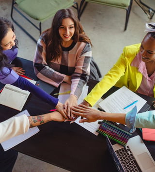 A group of diverse individuals sitting around a table, smiling and stacking their hands together in a gesture of teamwork and collaboration, with notebooks, documents, and a laptop visible on the table.