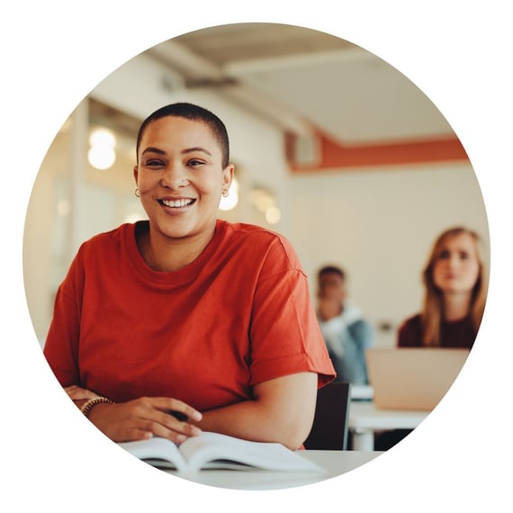 The image shows a person with short dark hair and a bright red t-shirt smiling broadly in a classroom setting. They are sitting at a table with an open book or notebook in front of them. The circular framing of the image creates a warm, engaging portrait in a professional or educational environment.
