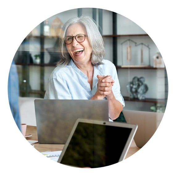 Smiling person with grey hair and glasses engaged in a conversation at a laptop during a meeting in a modern office space.