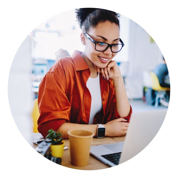 A person wearing glasses and an orange shirt is sitting at a desk, smiling while looking at a laptop screen. They have curly hair tied up and are resting their chin on one hand. A coffee cup and a small potted plant are on the desk.