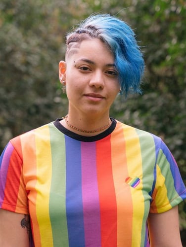 Person with short blue hair and shaved side patterns, wearing a rainbow-striped T-shirt with a small heart patch, standing outdoors in front of greenery.
