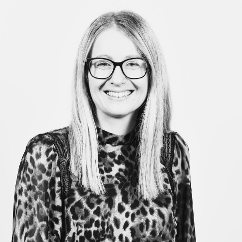 Grey scale image shows Janet Tidmarsh smiling at the camera. Janet has long hair and glasses. She is wearing an animal print blouse.